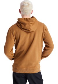 Persona con cabello corto y rubio claro, vistiendo un sudadera con capucha marrón y pantalones oscuros, vista desde atrás con las manos en las caderas.