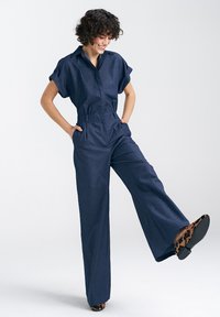Granatowy kombinezon z krótkimi, podwiniętymi rękawami, dopasowaną talią i szerokimi nogawkami. Wzorzyste buty z okrągłym czubkiem i niskim obcasem.