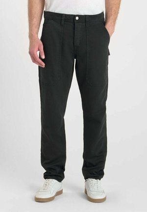 Man in zwarte casual broek met voorzakken en witte sneakers, staand met zijn handen in zijn zakken op een effen achtergrond.