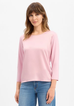 Jonge vrouw met lichtbruin haar, die een lichtroze top met lange mouwen en een blauwe spijkerbroek draagt, staand tegen een witte achtergrond.
