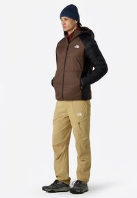 Giacca isolante marrone e nera con chiusura a zip, indossata sopra una camicia color vinaccia e abbinata a pantaloni kaki chiaro e scarpe da trekking nere.