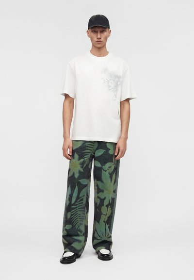 Άνδρας που φοράει μαύρο καπέλο, λευκό μπλουζάκι με ανοιχτό floral σχέδιο, φαρδιά παντελόνια με πράσινο φύλλωμα και ασπρόμαυρα παπούτσια, στέκεται όρθιος.