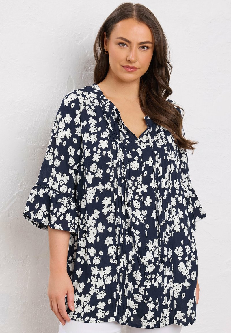 Marineblauwe bloemen tuniek met witte bloemenpatronen, v-hals en uitlopende mouwen. Gemaakt van lichte stof met geplooid ontwerp en relaxte pasvorm.