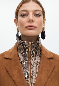 Hnědá semišová bunda s vysokým límcem se vzorem hada, zdobená zlatými flitry, zlatým zipem a velkými černými a zlatými náušnicemi.