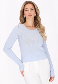 Maglione azzurro chiaro a maniche lunghe con scollo a scallop, caratterizzato da una texture a coste e un motivo perforato, indossato con pantaloni bianchi.