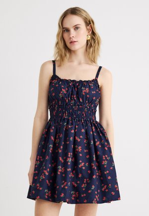 Νεαρή γυναίκα με κυματιστά ξανθά μαλλιά, που φοράει μπλε σκούρο αμάνικο φόρεμα με κόκκινο λουλουδάτο σχέδιο και λεπτομέρειες βολάν στο μπούστο, σε απλό φόντο.