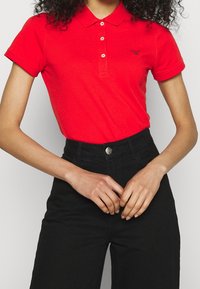 Femme portant un polo rouge ajusté avec des boutons et un pantalon noir taille haute, les mains jointes devant à hauteur de taille.