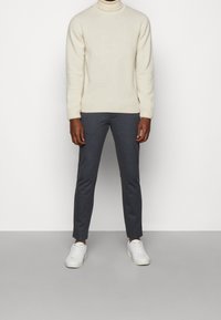 Maglione a collo alto color crema, pantaloni grigi aderenti e sneakers bianche. Il maglione ha una finitura testurizzata; i pantaloni presentano un motivo sottile.