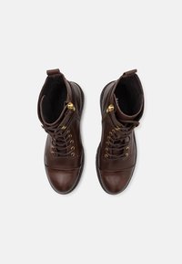 Une paire de bottes en cuir marron foncé à lacets avec des détails en fermeture éclair dorée, vues de dessus sur un fond blanc.