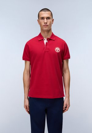 Hombre de pie mirando al frente, vistiendo una camisa tipo polo roja con un logo blanco en el pecho y pantalones azul oscuro, sobre un fondo liso.