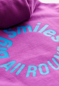 Lila Kapuzenpullover mit dem Aufdruck "Smiles All Round" in großen, fettgedruckten hellblauen Buchstaben, aus weichem Stoff mit strukturierter Oberfläche.