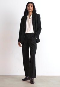 Costume noir à rayures, ajusté, associé à une chemise blanche à col. Le mannequin se tient avec assurance, portant des bottines noires.