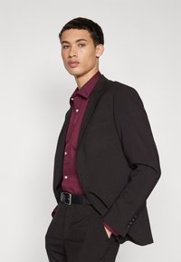 Chaqueta de traje negra sobre una camisa de botones burdeos, con una textura suave, un diseño ajustado y un simple cinturón negro que acentúa.
