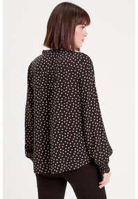 Black long-sleeve blouse with cream polka dots, gathered cuffs, and a subtle ruffle at the neckline; relaxed fit and smooth texture.