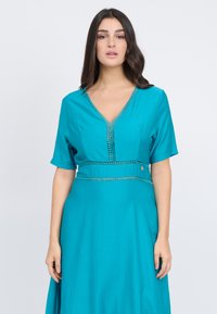 Türkises Kleid aus strukturiertem Stoff. V-Ausschnitt mit dekorativer Stickerei und kurzen Ärmeln, in der Taille mit einem flachen Band gerafft.