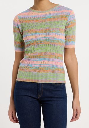 Vrouw die een kortärmelig, meerkleurig pastelkleurig gestreept kabelgebreid trui draagt en donkerblauwe spijkerbroek, staand voor een witte achtergrond.