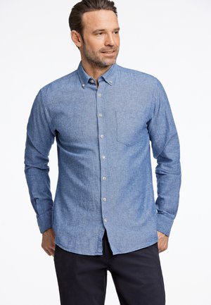 Chemise bleu clair à manches longues en tissu texturé, dotée d'une poche, d'un ourlet arrondi et d'un col boutonné. Portée avec un pantalon sombre.