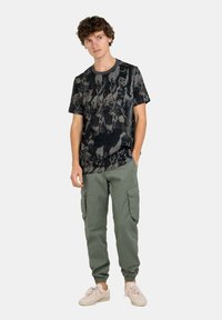 Camiseta negra de manga corta con un patrón abstracto en gris, combinada con pantalones cargo verdes claros. El modelo lleva zapatillas beige; fondo neutro.