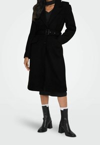 Manteau en laine noire avec taille ceinturée, col cranté et poches latérales. Porté sur une robe sombre avec des bottines noires et des socquettes blanches.