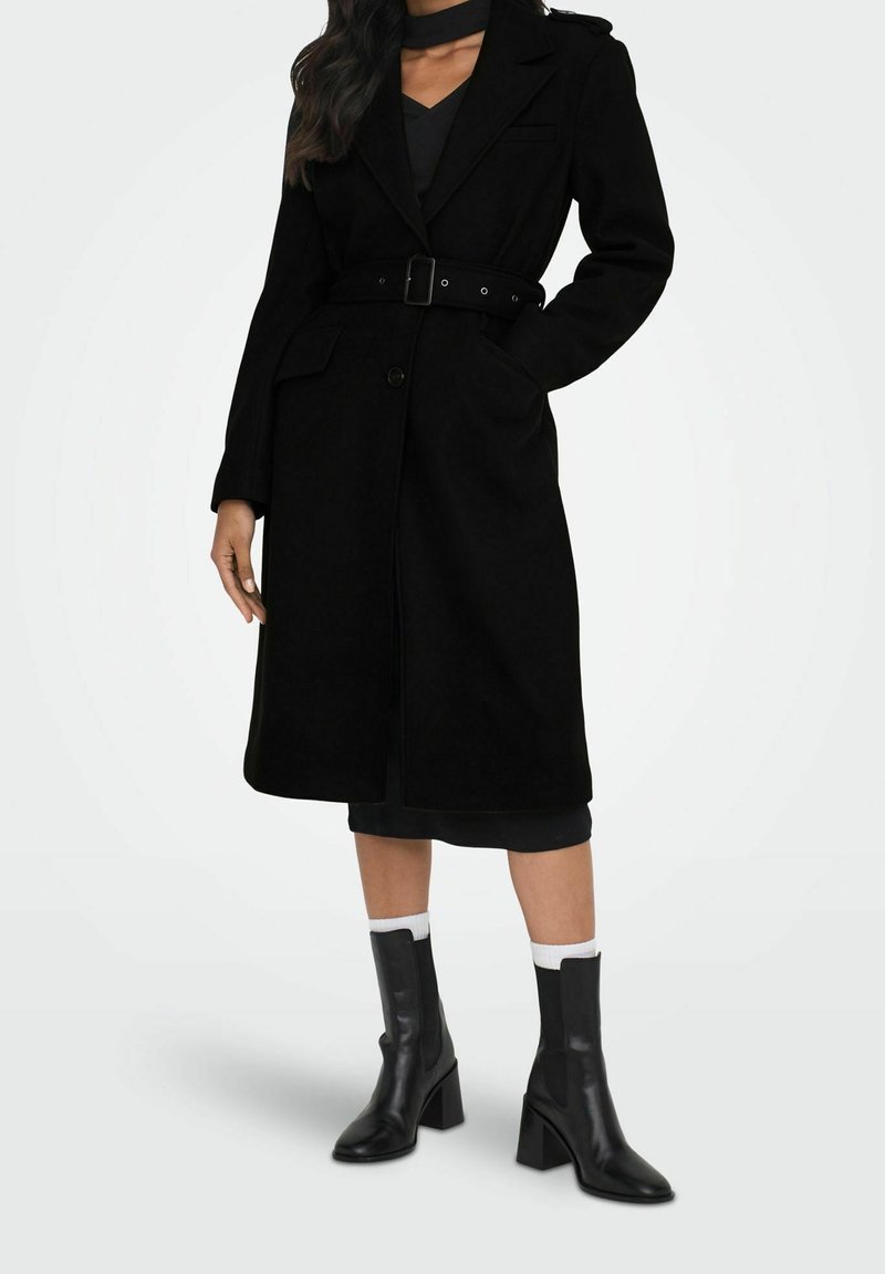 Manteau en laine noire avec taille ceinturée, col cranté et poches latérales. Porté sur une robe sombre avec des bottines noires et des socquettes blanches.