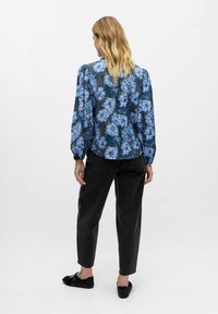 Blusa floreale blu e bianca su uno sfondo scuro, con maniche a palloncino e orlo arrotondato. Abbinata a pantaloni neri slim e scarpe nere.