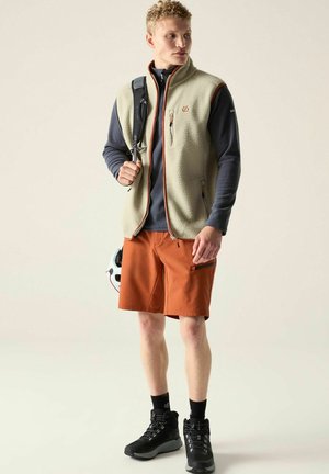 Young man wearing beige fleece vest, navy long-sleeve shirt, orange shorts, black hiking boots, carrying a backpack and holding a white helmet.
