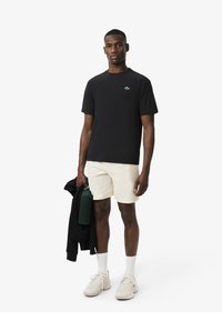 Camiseta negra de manga corta con un pequeño logo verde, pantalones cortos beige claro, calcetines blancos y zapatillas beige, sosteniendo una botella de agua verde y una chaqueta.