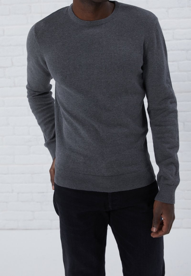 Homme portant un pull gris foncé ajusté à col rond et un pantalon noir, debout devant un mur en briques blanches.