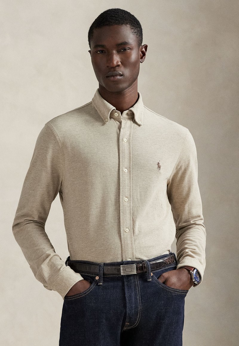 Camisa caqui de mangas compridas com botões e gola. Apresenta um pequeno logótipo no peito, combinada com jeans escuros e um cinto.