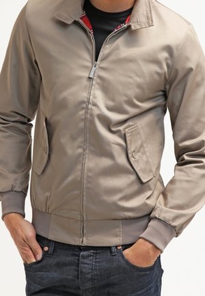 Mand iført beige lynlåsjakke med knaplukket klaplommer og ribstrikkede manchetter, hænderne i lommerne, kombineret med mørkeblå jeans.