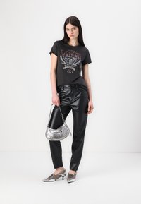 Grijze graphic t-shirt met een adelaarontwerp, zwarte leren broeken, zilveren handtas en metallic schoenen met kleine hakken. Effen witte achtergrond.