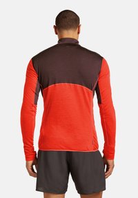 Hombre vestido con una camiseta atlética de manga larga roja y marrón oscuro y pantalones cortos negros, de pie de espaldas frente a un fondo liso.
