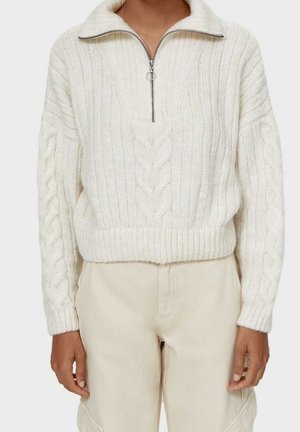 Personne portant un pull en maille torsadée crème avec un col à demi-fermeture éclair et un pantalon beige clair assorti, debout devant un fond uni.