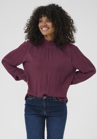 Maroon blouse with long sleeves, gathered neckline, and textured fabric. Paired with dark blue fitted jeans. Simple and elegant design.