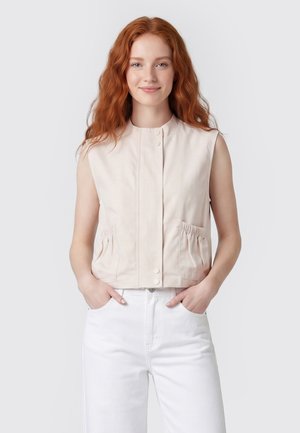 Ung kvinde med langt rødt hår iført en ærmeløs beige vest og hvide bukser, stående med hænderne i lommerne mod en ensfarvet baggrund.