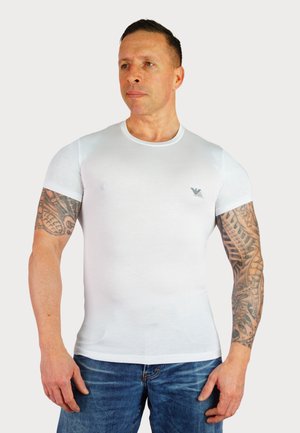 Weißes T-Shirt mit kurzen Ärmeln aus Baumwolle, figurbetontes Design, glatte Textur, mit einem kleinen Logo auf der linken Brust, getragen mit blauen Jeans.