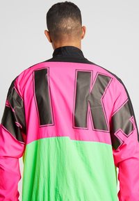 Man som bär en neongrön och pink jacka med stora svarta blockbokstäver på ryggen, står vänd bort från kameran mot en enkel bakgrund.