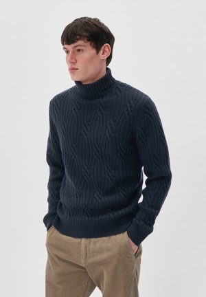 Granatowy sweter z golfem, dziany, z teksturowanym wzorem i ściągaczami na rękawach. Noszony z jasnobrązowymi spodniami z korduroju.