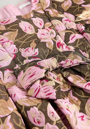 Textilie s květinovou výšivkou v odstínech růžové a béžové na hnědém pozadí, s texturovaným vzorem a detailem mašle.