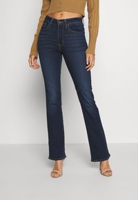 Mörkblå utsvängda jeans med midjehöjd, femficksdesign och kontrasterande sömmar, tillsammans med beige klacksandaler.