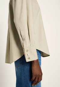 Beige long-sleeve shirt cuff with four buttons, worn over blue denim jeans, showing side view of arm and hand against a neutral background.