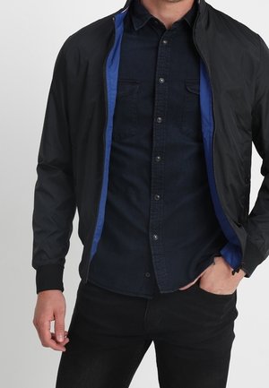 Un homme porte une veste noire à fermeture éclair avec une doublure intérieure bleue par-dessus une chemise sombre à boutons et un pantalon noir, une main dans la poche, sur fond blanc.