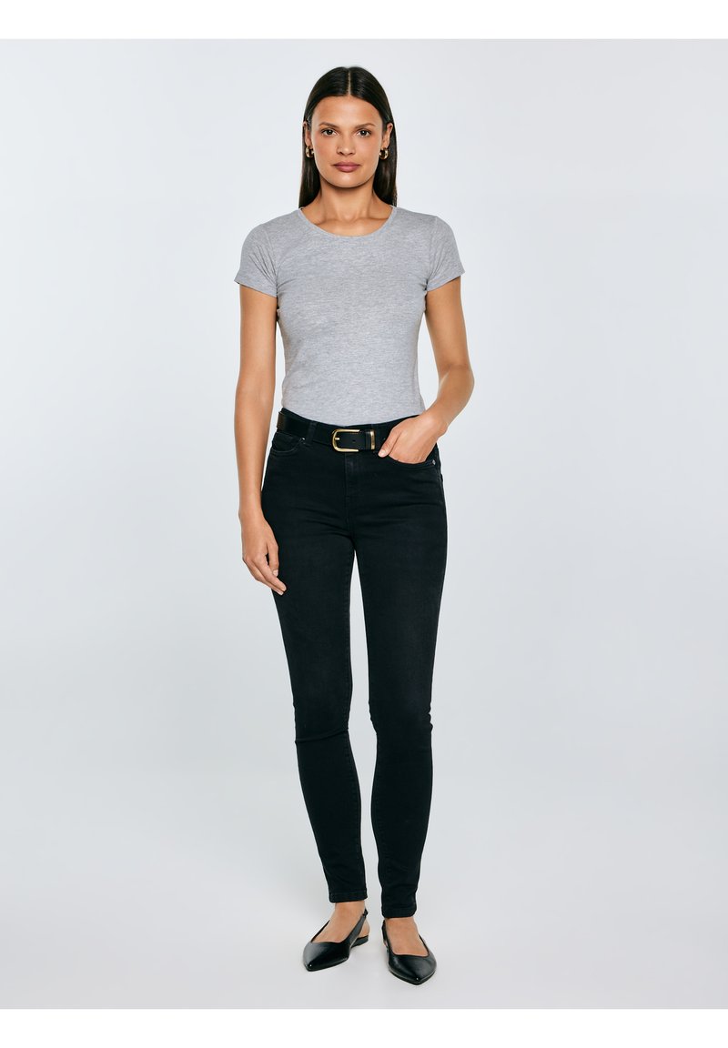 Kobieta stojąca prosto, ubrana w szarą koszulkę z krótkim rękawem, czarne dżinsy z paskiem, czarne baleriny oraz złote kolczyki koła.