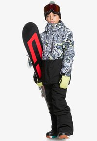Enfant portant une veste de neige à motifs avec une base noire, un pantalon noir et des gants vert citron, tenant un snowboard avec des accents rouges. Bottes marron.