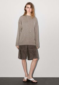 Beiger oversized Pullover, schokoladenbraune Wildledershorts, weiße Flip-Flops. Lange Ärmel, lockere Passform, Anhänger-Halskette, glatte Textur.