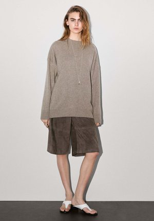 Maglione oversize beige, pantaloni corti in suede marrone, infradito bianche. Maniche lunghe, vestibilità rilassata, collana con ciondolo, texture liscia.
