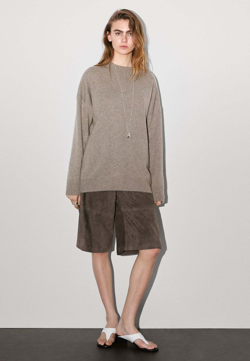 Beiger oversized Pullover, schokoladenbraune Wildledershorts, weiße Flip-Flops. Lange Ärmel, lockere Passform, Anhänger-Halskette, glatte Textur.