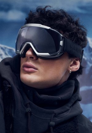 Slidinėjimo akiniai su tamsiais lęšiais ir sidabro akcentais; minkšta juoda elastinė juosta su „HAWK“ logotipu; dėvimi asmens su garbanotais plaukais ir džemperiu su gobtuvu.