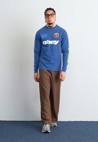 Camisa de manga larga azul con texto blanco "obey" y detalles verdes. Pantalones de cuadros marrones. Zapatillas grises. Fondo liso, modelo de pie.