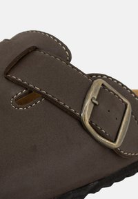 Sandale en suède marron avec une sangle réglable et une boucle métallique ; présente une couture beige et une semelle texturée et flexible.